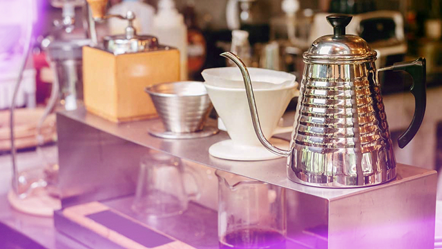 Close-up of a coffee brewing setup featuring a reflective stainless steel kettle, ceramic drippers, and glass servers on a counter, with a cozy café vibe